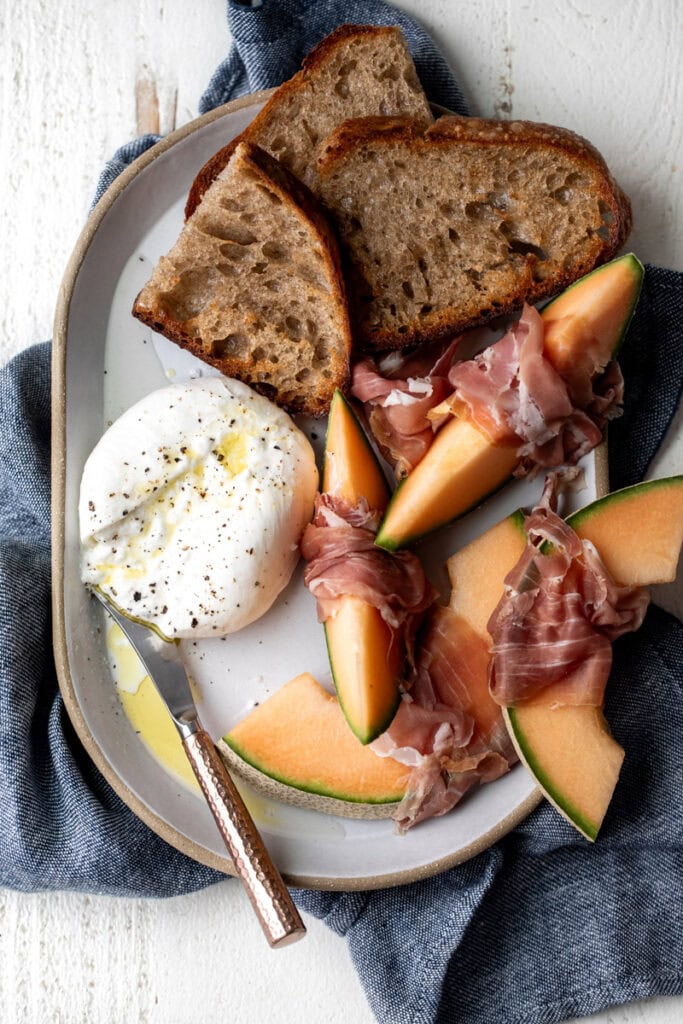 burrata and prosciutto snack plate with cantaloupe and toasted garlic bread on platter.