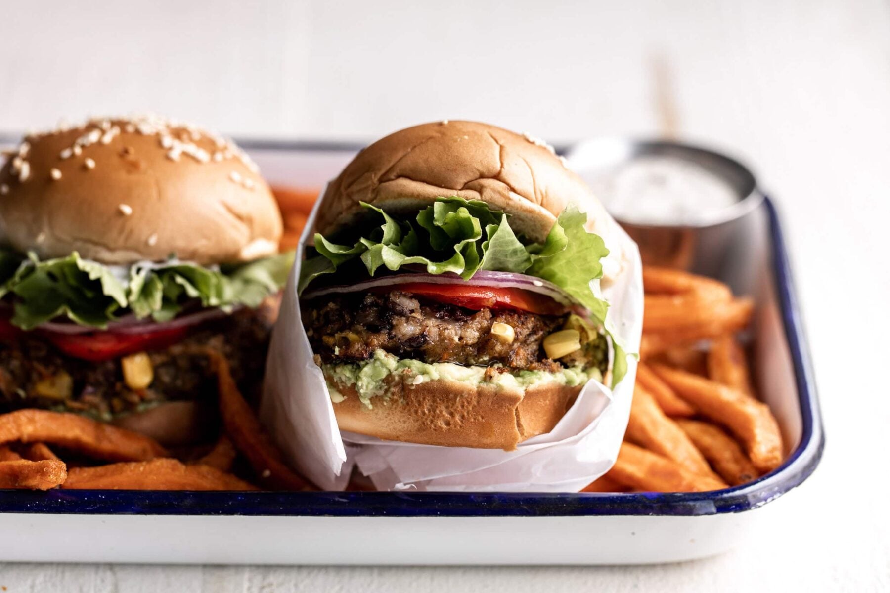 spicy black bean and corn veggie burgers served with tomato, onion, lettuce on hamburger buns wrapped in white parchment paper on a tray with sweet potato fries