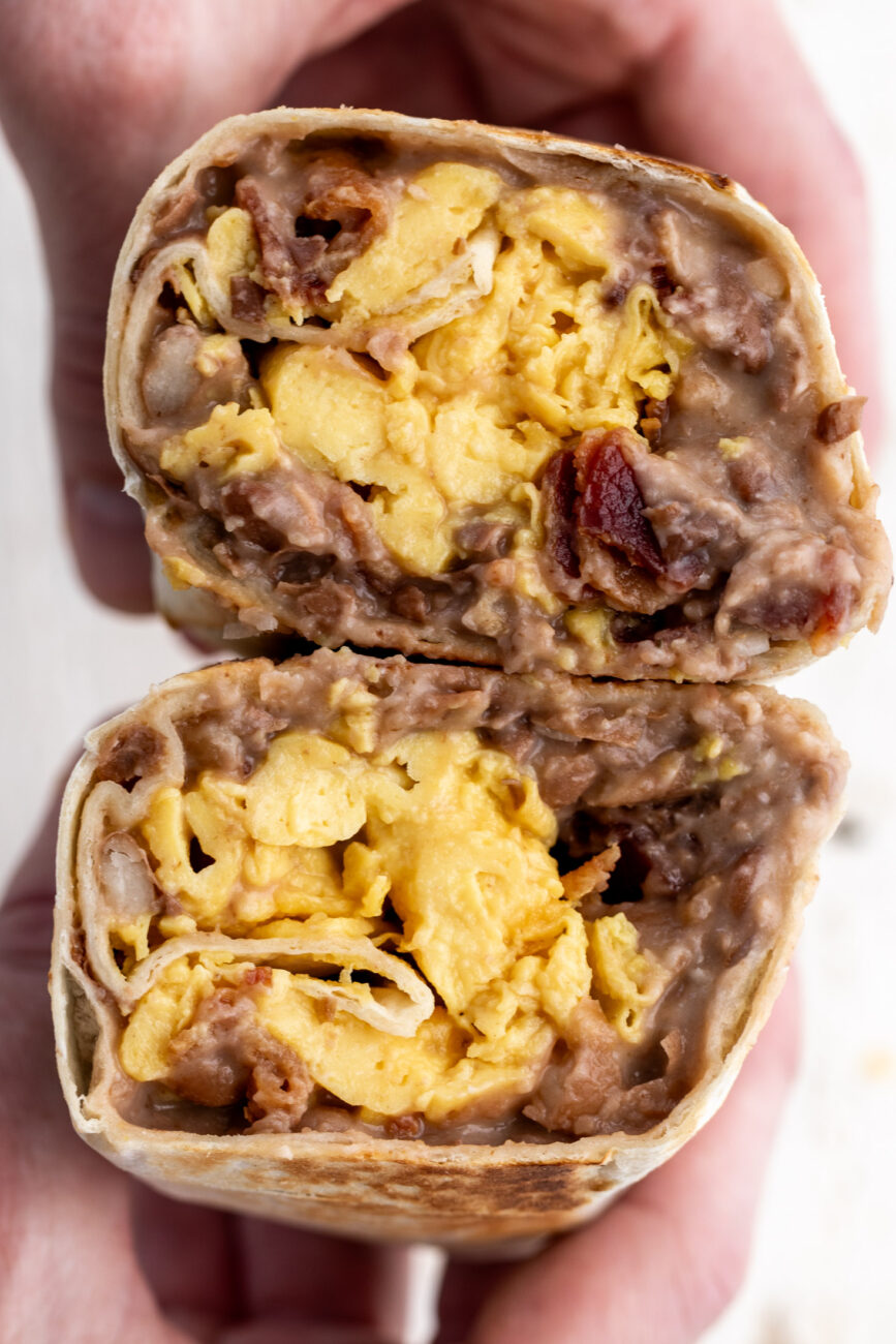 bacon breakfast burrito with hot sauce dripping onto bean, cheese egg and bacon filling cut open.
