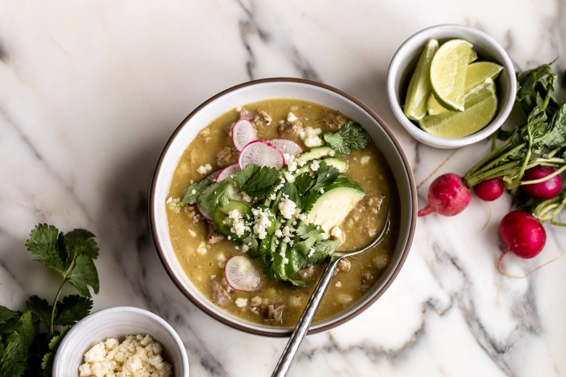 pork pozole verde with sliced avocado, radish, cilantro, jalapeño slices. Lime wedges on the side.