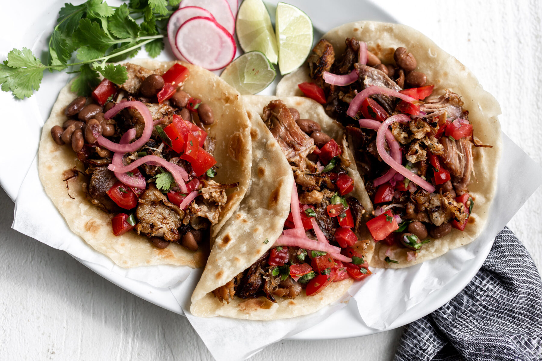 shredded pork carnitas on flour tortillas with pickled onions and tomatoes on a white plate with blue napkin.