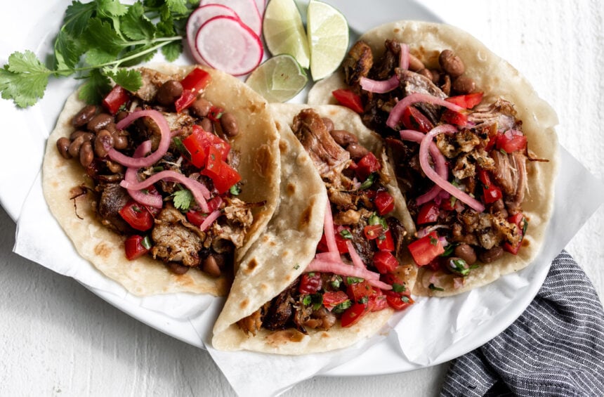 shredded pork carnitas on flour tortillas with pickled onions and tomatoes on a white plate with blue napkin.