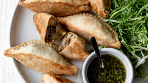 Beef, onion and potato empanadas with chimichurri sauce on a platter. One Empanada cut in half to show the center.