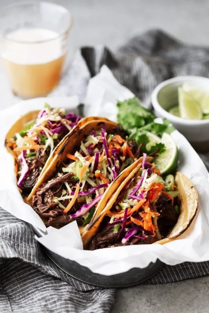 three Braised Korean BBQ Beef Tacos with broccoli slaw on parchment paper.