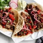 shredded pork carnitas on flour tortillas with pickled onions and tomatoes on a white plate with blue napkin