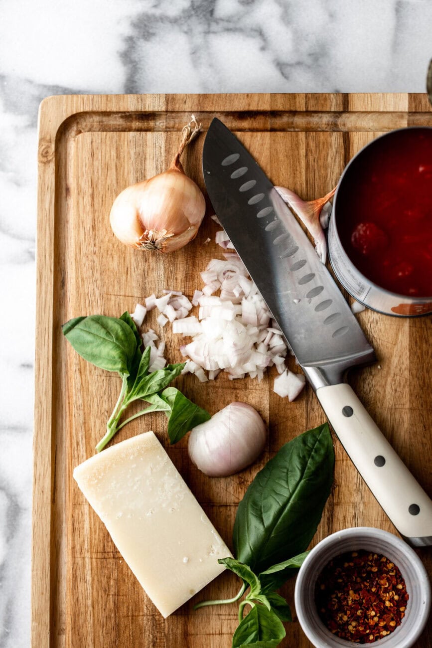 Overhead ingredient shot of a diced onion with chefs knife. Also on the cutting board is a can of tomatoes, block of cheese, and basil.