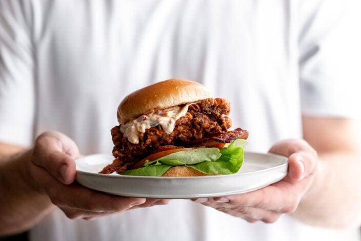 Pimento cheese fried chicken sandwich with lettuce and tomato on a plate being held.