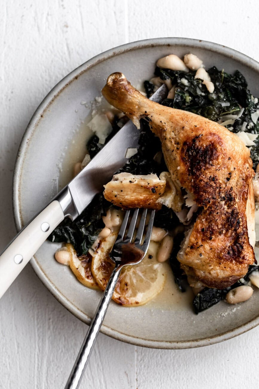 Roast Chicken Legs with Braised Kale and White Beans with parmesan cheese and lemons cut with fork and knife.