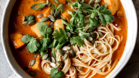 chunks of pumpkin in a red coconut curry broth with thin noodles in a white bowl topped with pumpkin seeds and cilantro