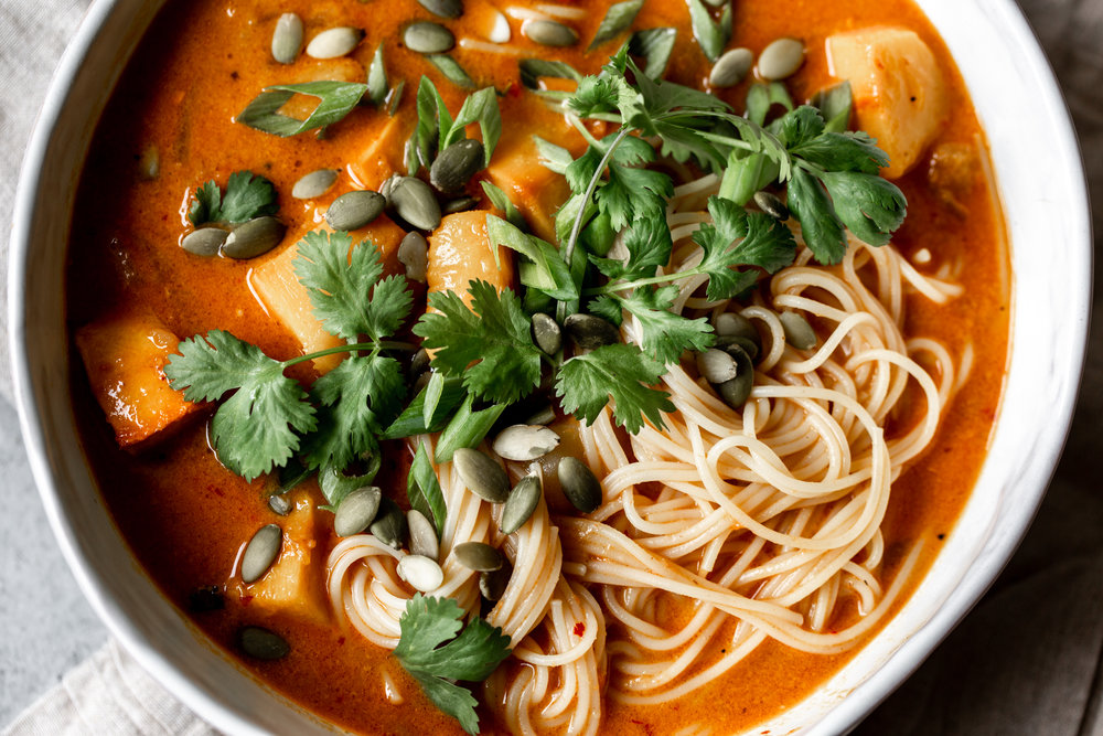 chunks of pumpkin in a red coconut curry broth with thin noodles in a white bowl topped with pumpkin seeds and cilantro
