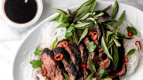 soy lime marinated beef seared and sliced over bean thread noodles with limes and thai basil leaves on a white plate and marble background