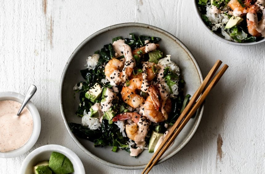marinated kale and crispy fried rice topped with green onions spicy mayo and avocado in a grey stone bowl with wood chopsticks on a white wood board.