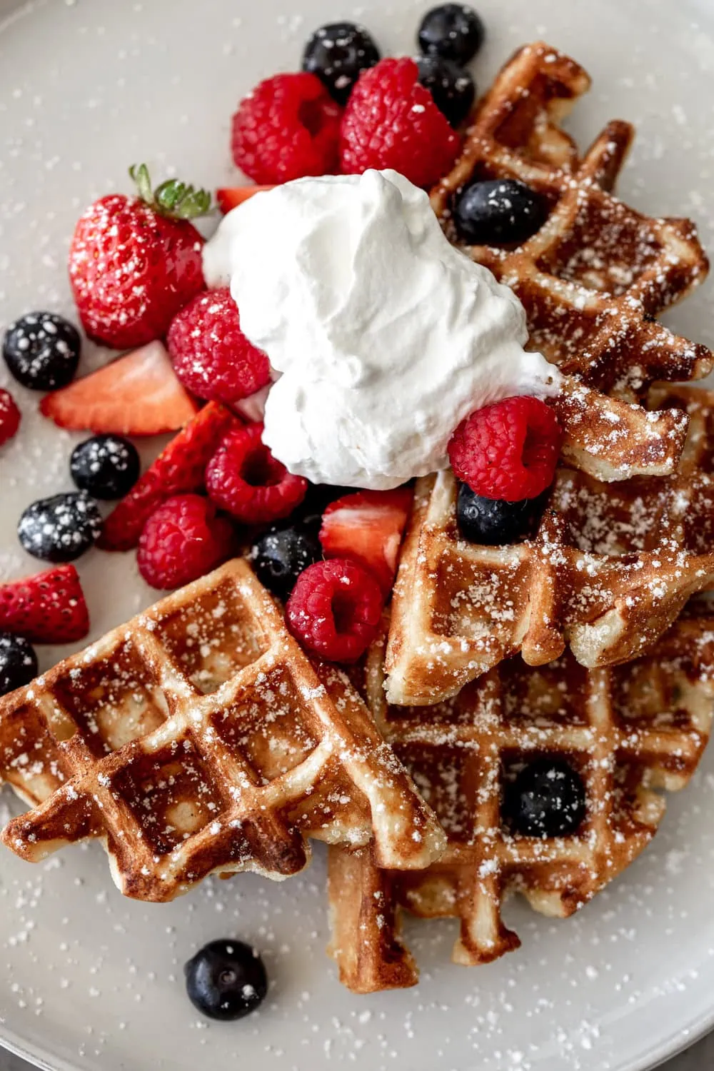 Yeasty Overnight Waffles topped with whipped cream strawberries raspberries and blueberries with powdered sugar