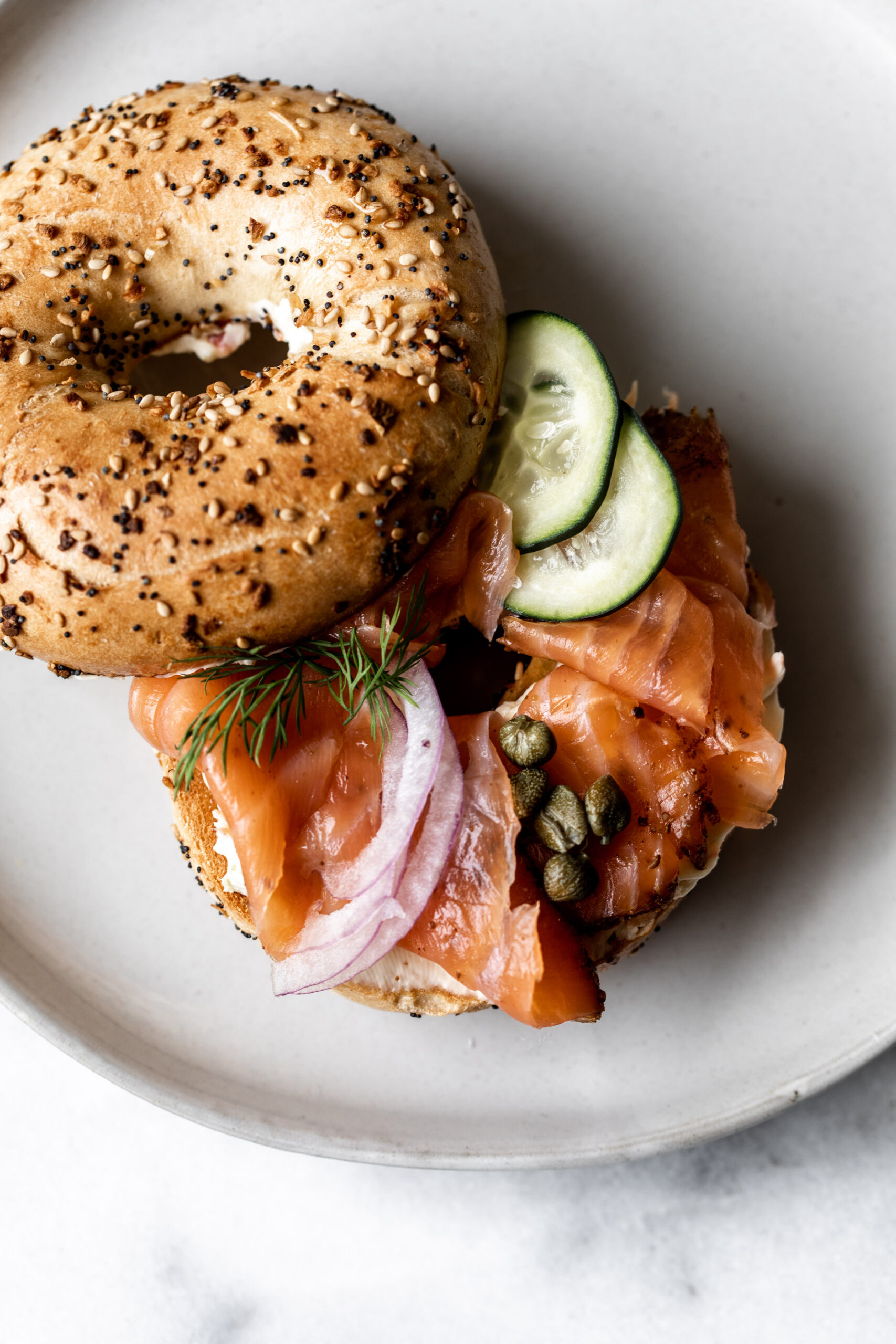 smoked salmon lox on an everything bagel with cucumber, capers, red onion on a white plate on marble