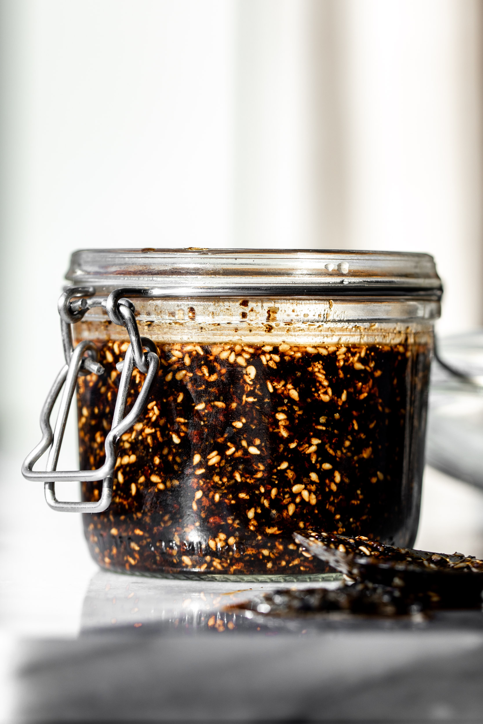 blended chilies, sesame seeds in oil in a small glass jar on a marble table in direct sunlight