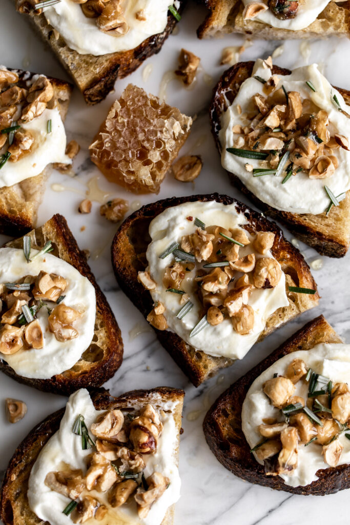 toasts topped with whipped ricotta, candied hazelnuts and rosemary on a white marble surface with honeycomb