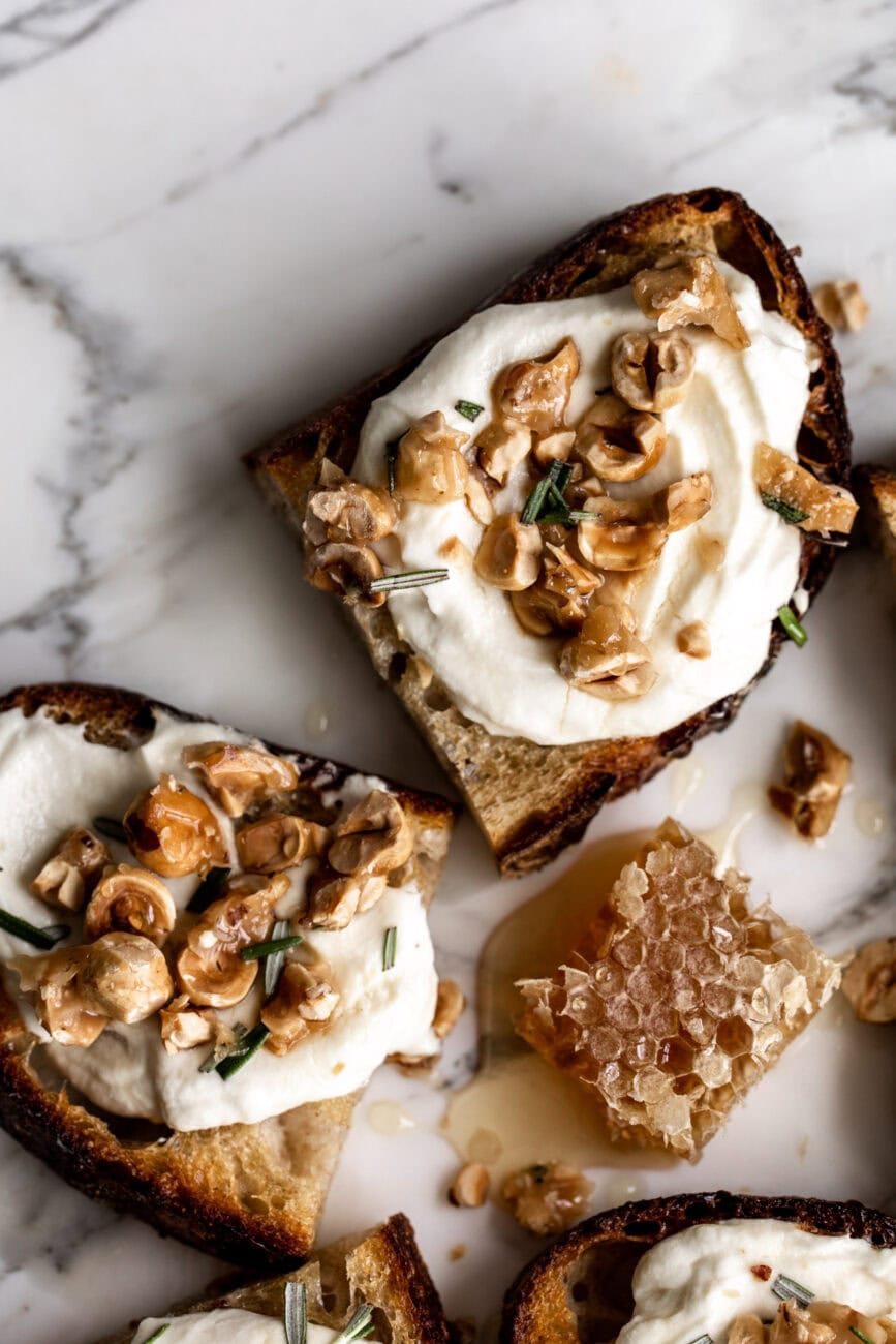 one toasts topped with whipped ricotta, candied hazelnuts and rosemary on a white marble surface with honeycomb.