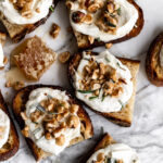 toasts topped with whipped ricotta cheese, toasted hazelnut brittle and rosemary with honey on white marble surface