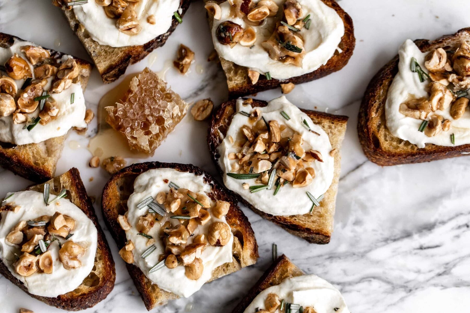 toasts topped with whipped ricotta cheese, toasted hazelnut brittle and rosemary with honey on white marble surface.
