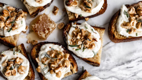 toasts topped with whipped ricotta cheese, toasted hazelnut brittle and rosemary with honey on white marble surface