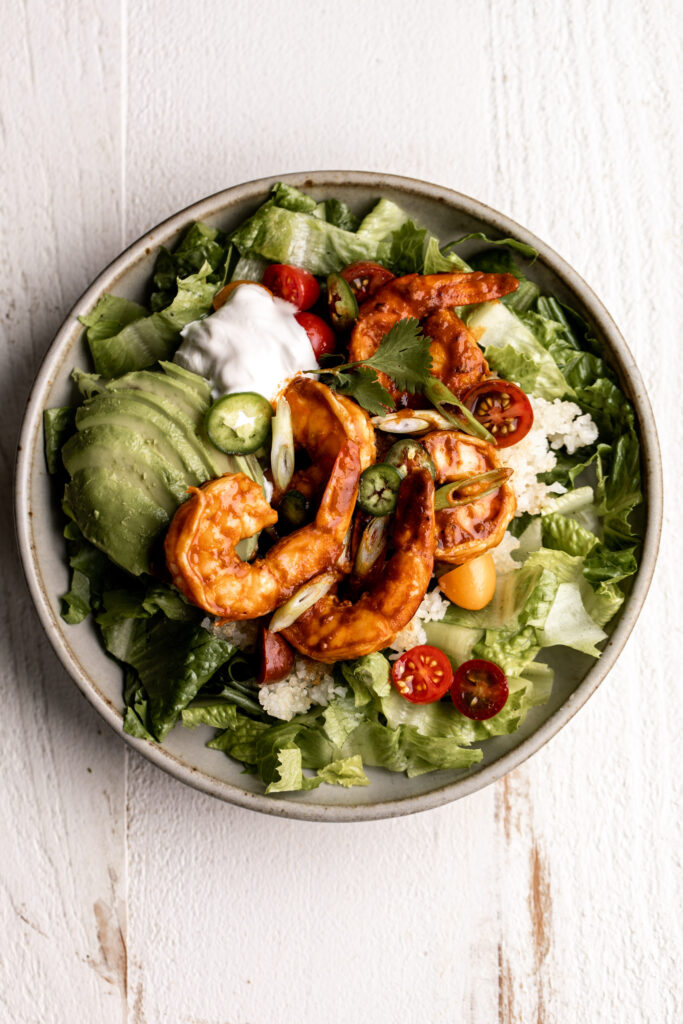 spicy shrimp burrito bowl with crispy rice, romaine lettuce, tomatoes, sour cream and avocado in a ceramic bowl