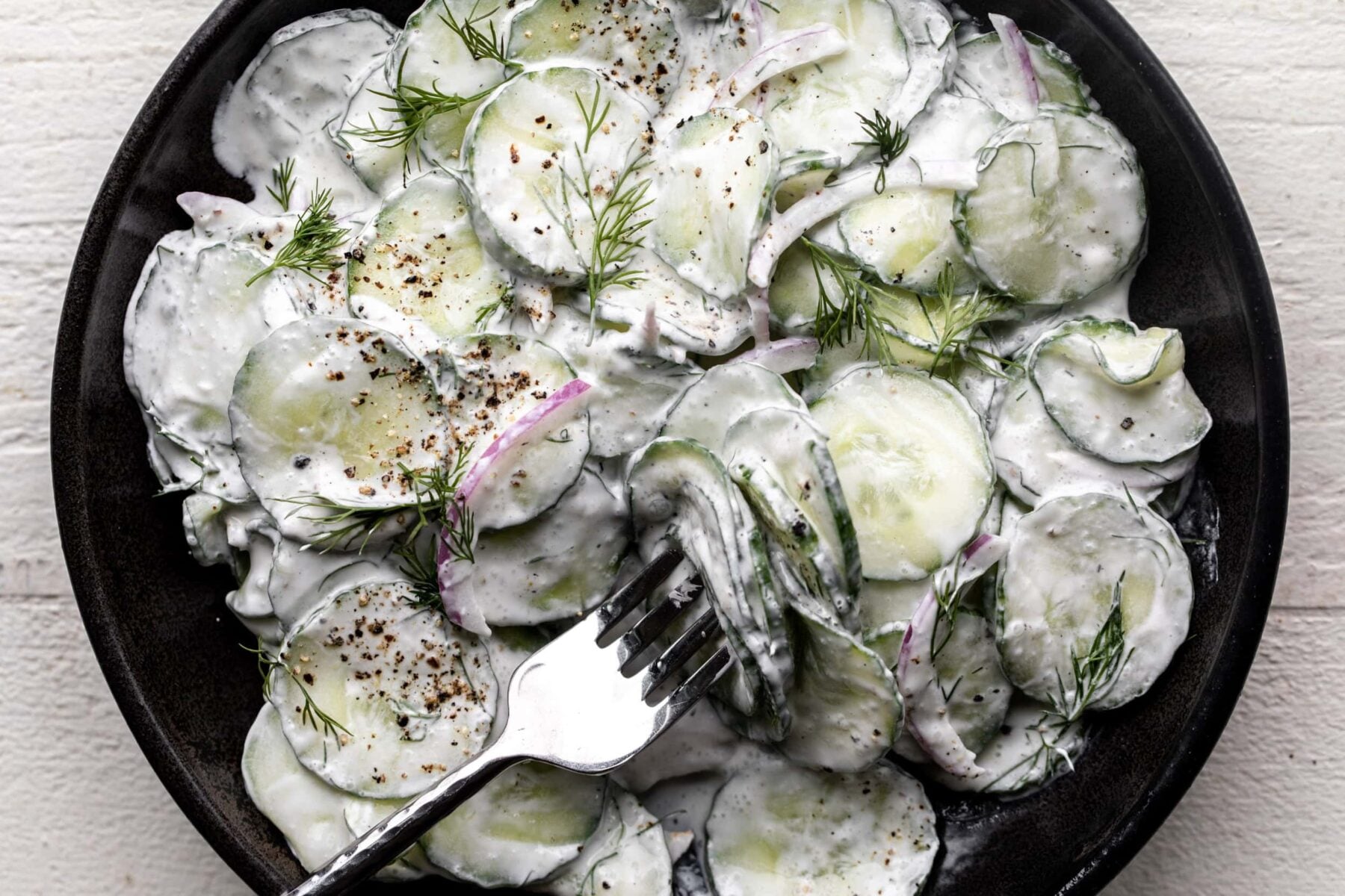 creamy cucumber salad with sour cream sauce, dill and red onion in black stone bowl.