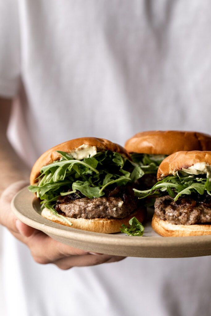 holding plate of lamb burgers made on with caramelized onions, arugula and curry mayo