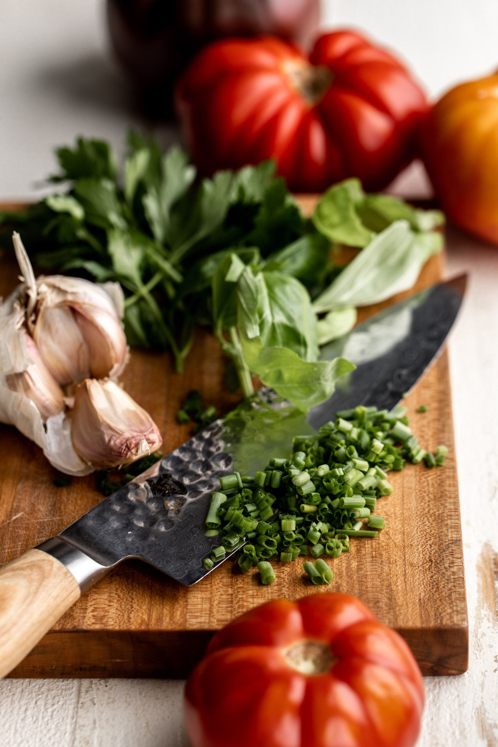 chopped chives with basil, heirloom tomato, basil and parsley on a small cutting board with a knife.