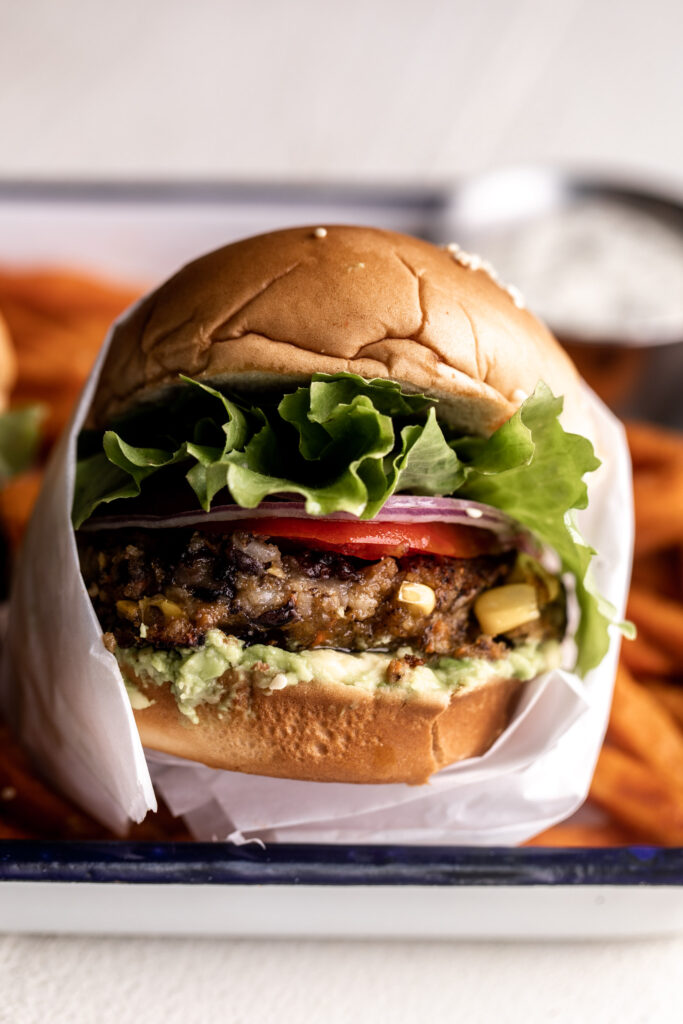 spicy black bean and corn veggie burgers served with tomato, onion, lettuce on hamburger buns wrapped in white parchment paper on a tray with sweet potato fries closeup photo