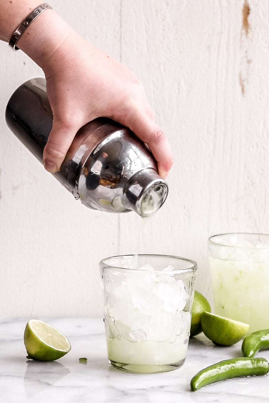 pouring margarita from cocktail shaker to glass with ice.