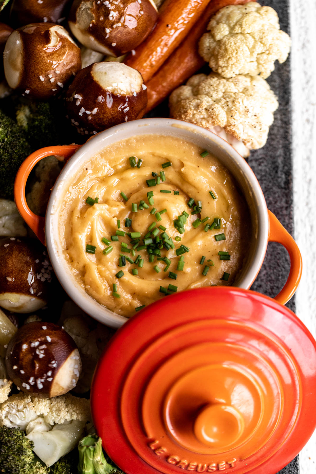 closeup cheddar beer cheese fondue in le creucet pot with roasted vegetables and pretzel balls.