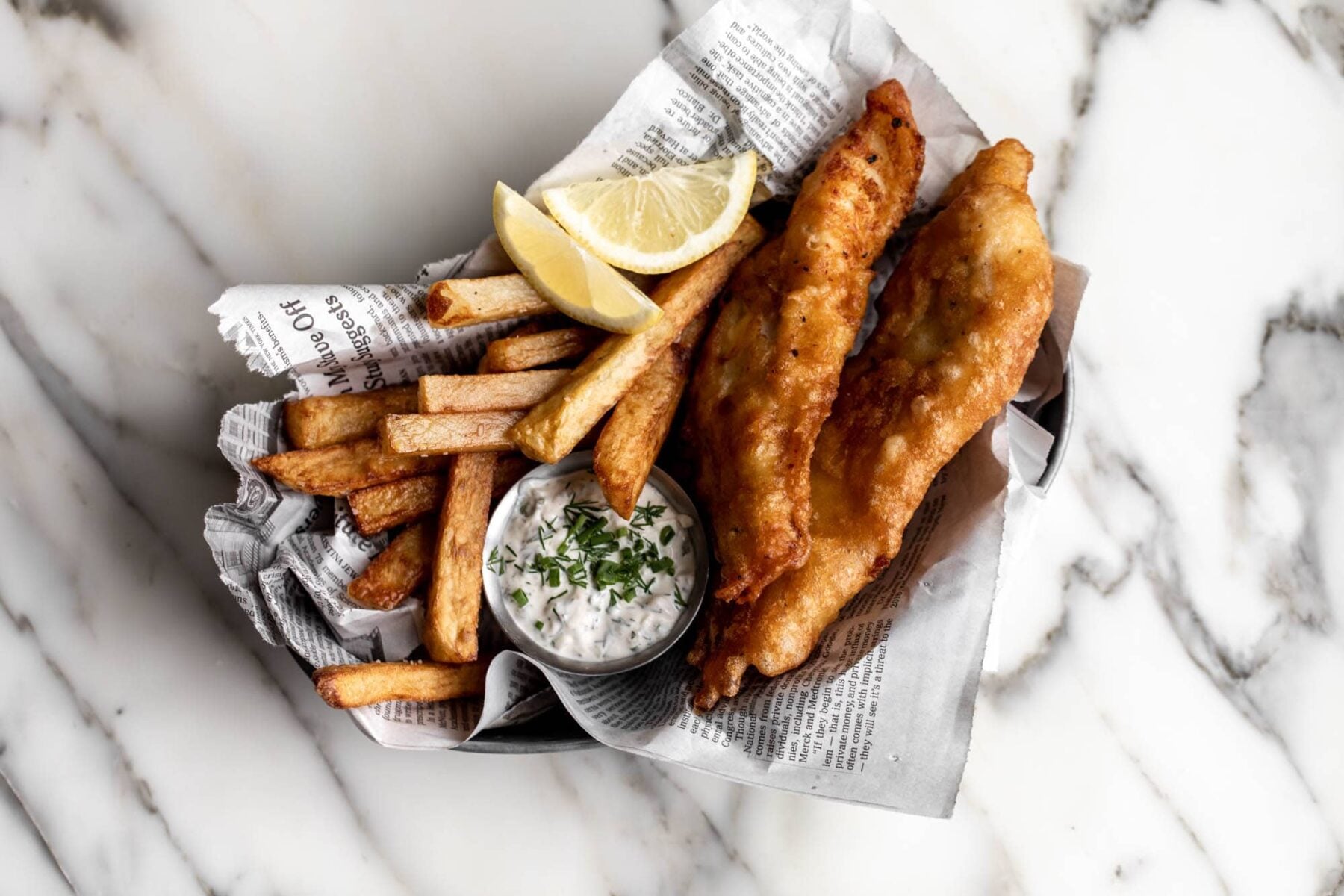 beer battered fried fish and chips with tartar sauce served in newspaper with a lemon wedge.