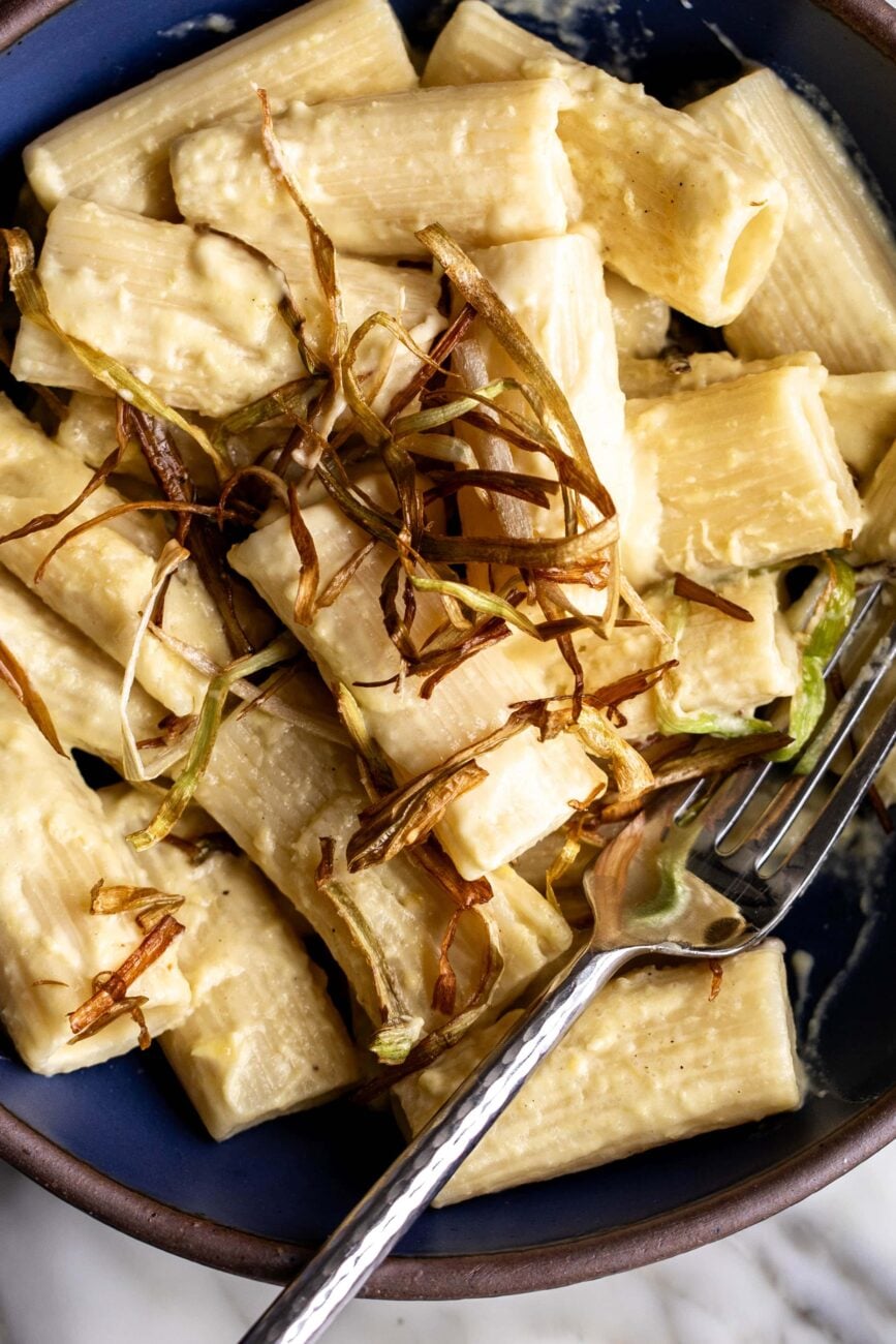 creamy corn rigatoni pasta topped with frizzled leeks and truffle oil with fork in blue stone bowl.