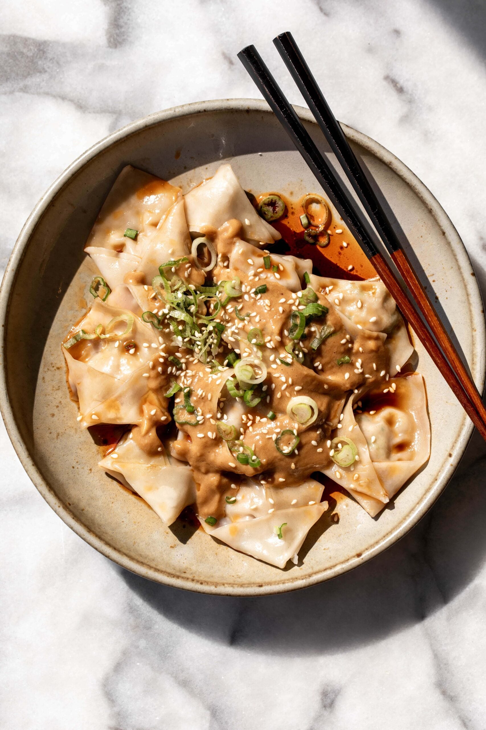 pork wontons with peanut sauce and chili oil garnished with chives and sesame seeds.