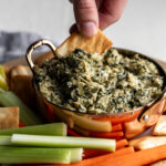 dipping pita chip in creamy spinach artichoke dip with celery and carrots.