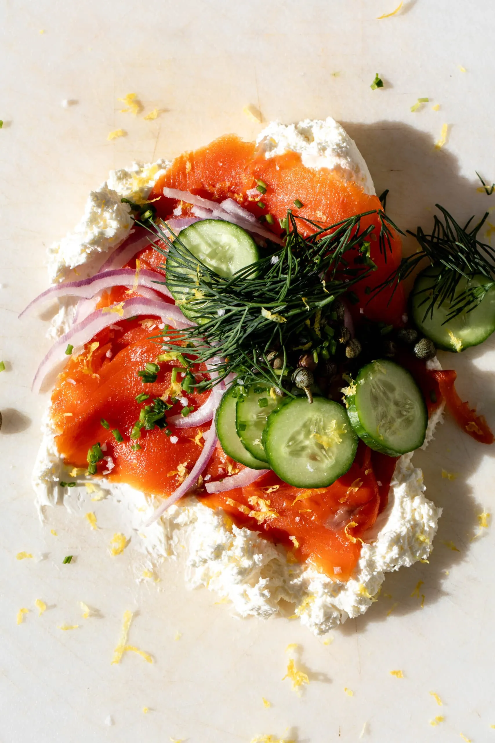 cream cheese on cutting board piled with smoked salmon, cucumber, dill and onion.