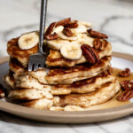 best banana pancakes stacked on plate with sliced bananas, pecans and maple syrup.