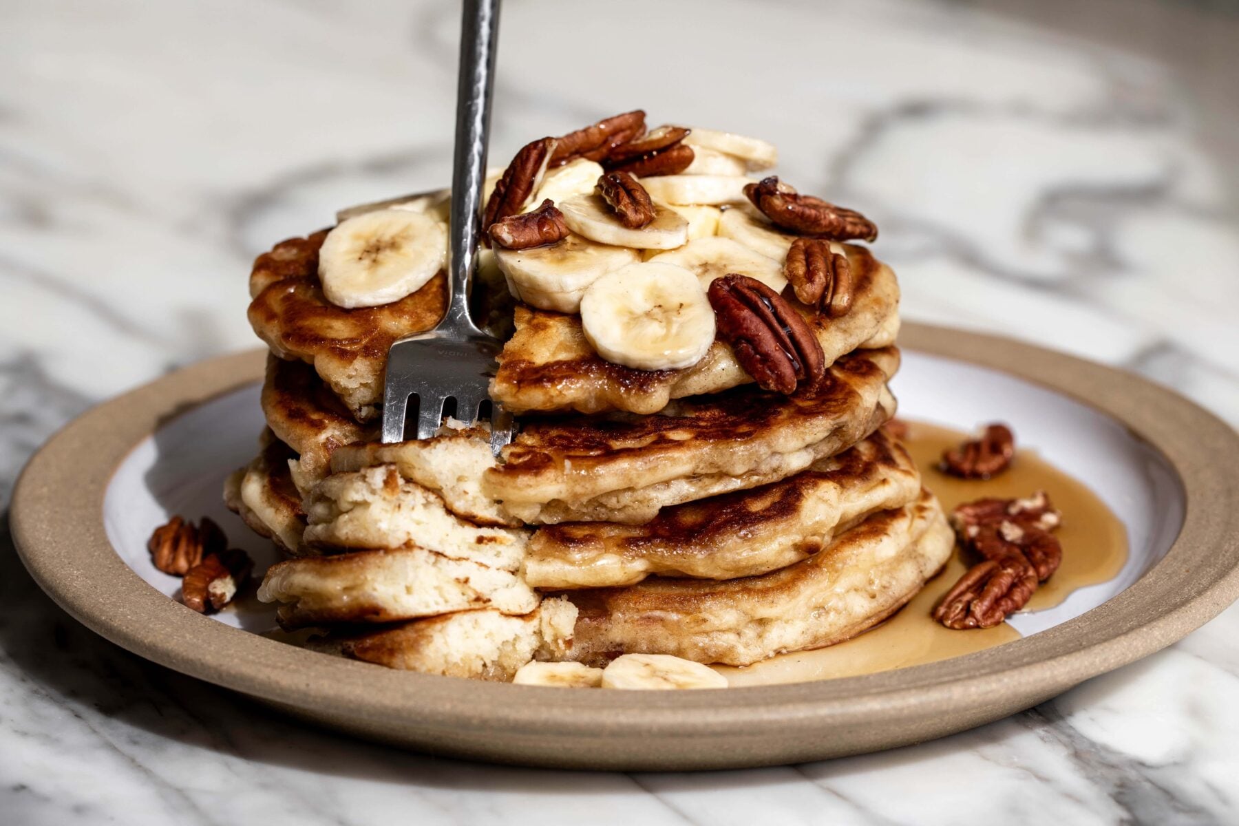 best banana pancakes stacked on plate with sliced bananas, pecans and maple syrup.