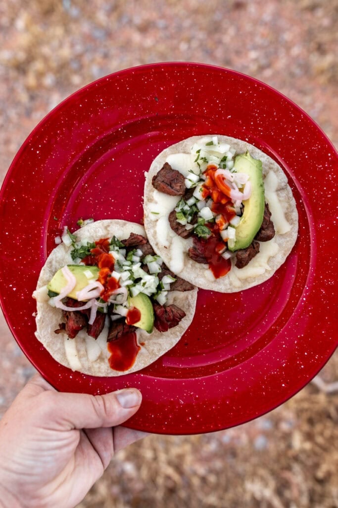 seared steak tacos with avocado slices and hot sauce on red plate.