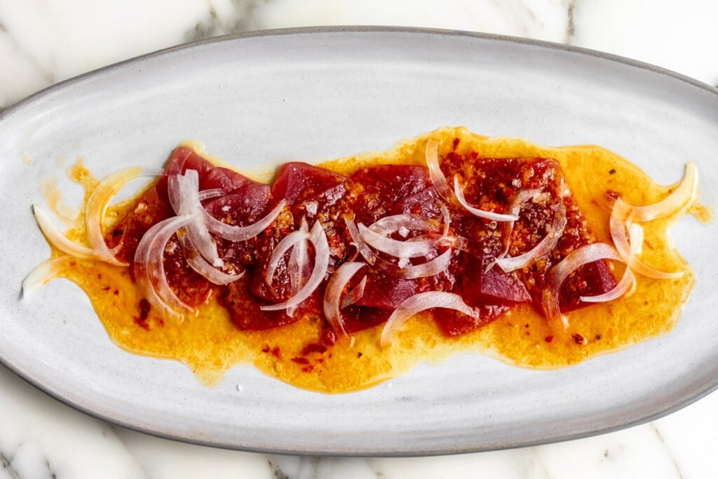 orange calabrian chili tuna crudo topped with shaved onion on oval plate.