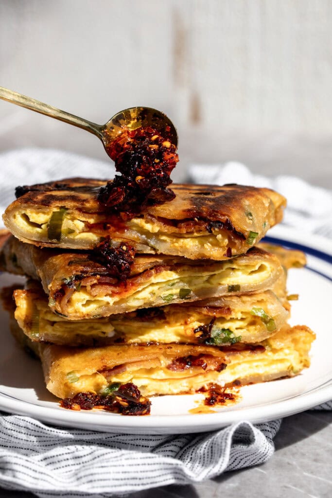 BEC scallion pancake breakfast sandwich stacked on plate with spoonful of chili crisp.