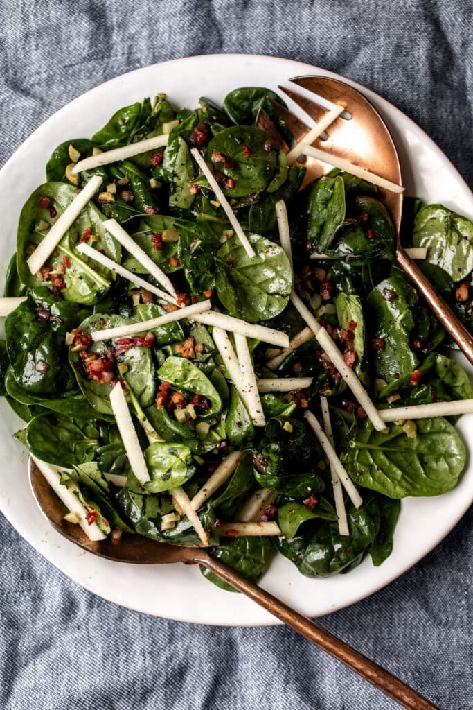 Warm Spinach Salad with copper serving spoons topped with pancetta and apples on denim napkin.