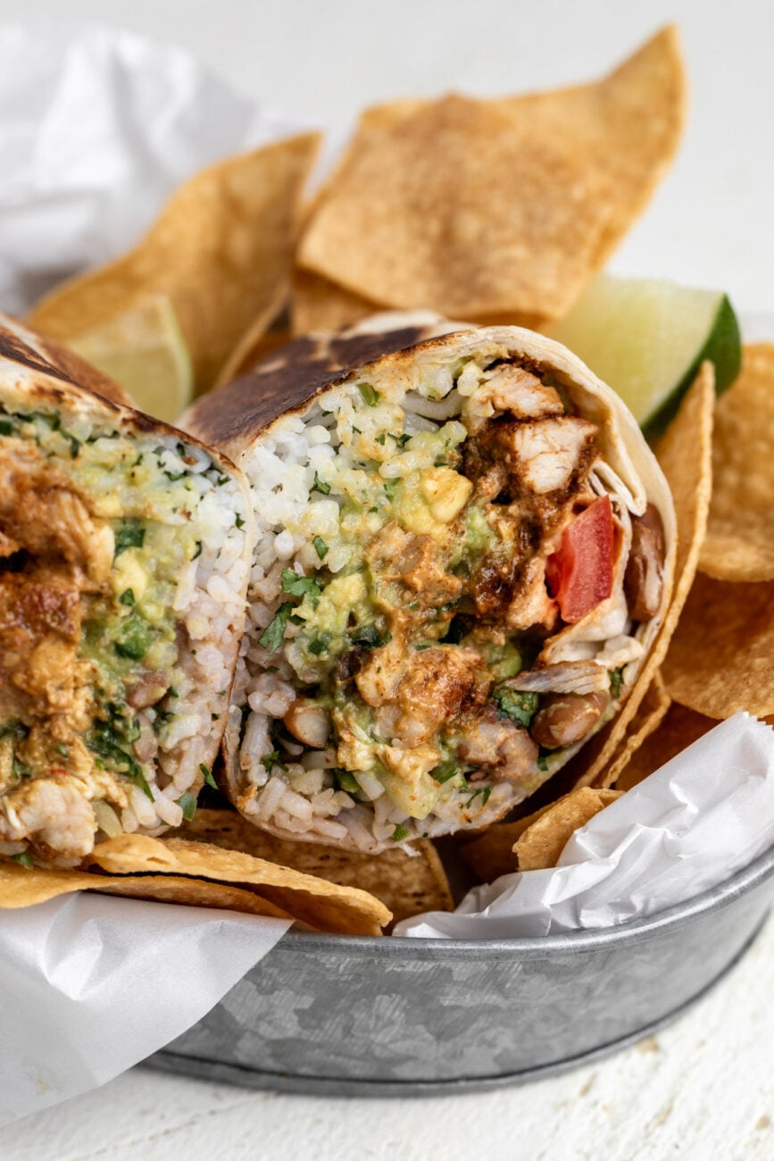 creamy chipotle chicken burrito cut in half with seasoned chicken pinto beans and rice with a side of chips.